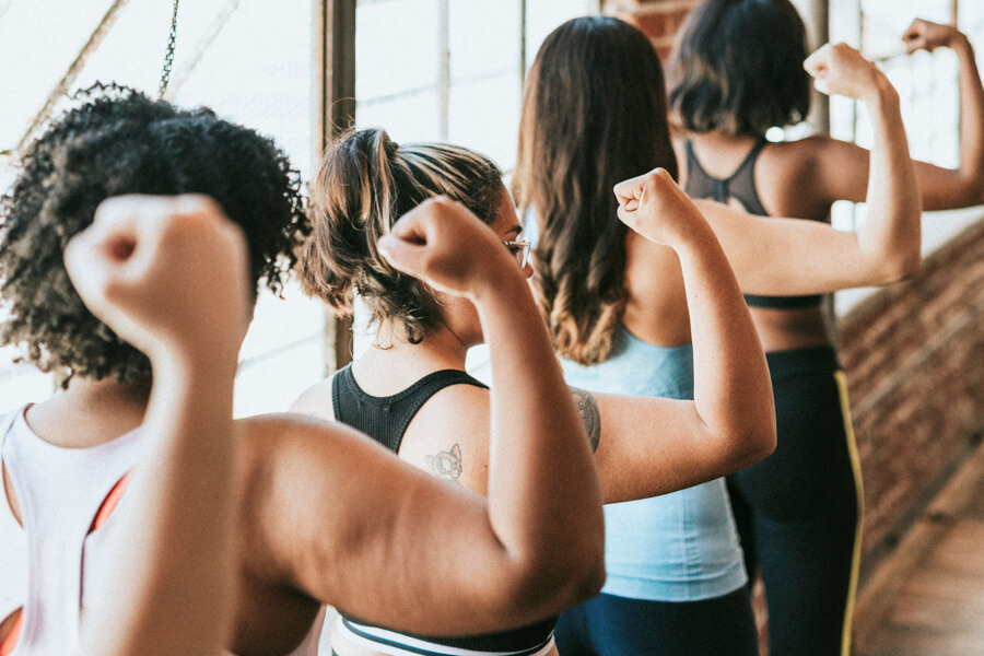 A group of diverse women in workout clothes flexing their arms and backs to the camera, symbolizing female strength, fitness, and encouraging movement.