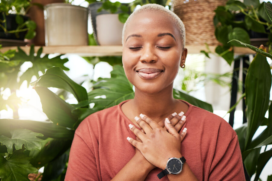 A woman with short hair standing among green plants, holding her hands over her heart with her eyes closed, symbolizing inner peace, spiritual grounding, and faith.